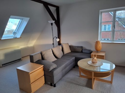 Livingroom 1 Heller Wohnraum mit grauem Sofa, Couchtisch und zwei Holzmöbeln am Fenster.